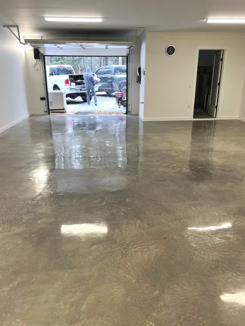 Empty garage with polished concrete floor, open garage door, person outside in a hoodie, two pickup trucks parked outside, and some trees in the background.