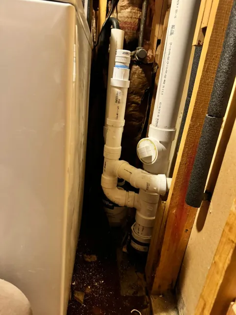 White PVC pipes and a valve in a narrow utility space beside a washer.