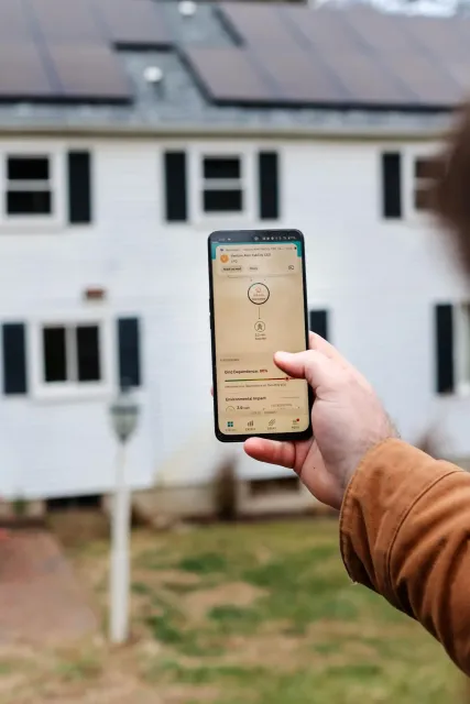 A person using a phone, the screen showcases a home automation app interface connected to smart electrical panels. The background is softly blurred.