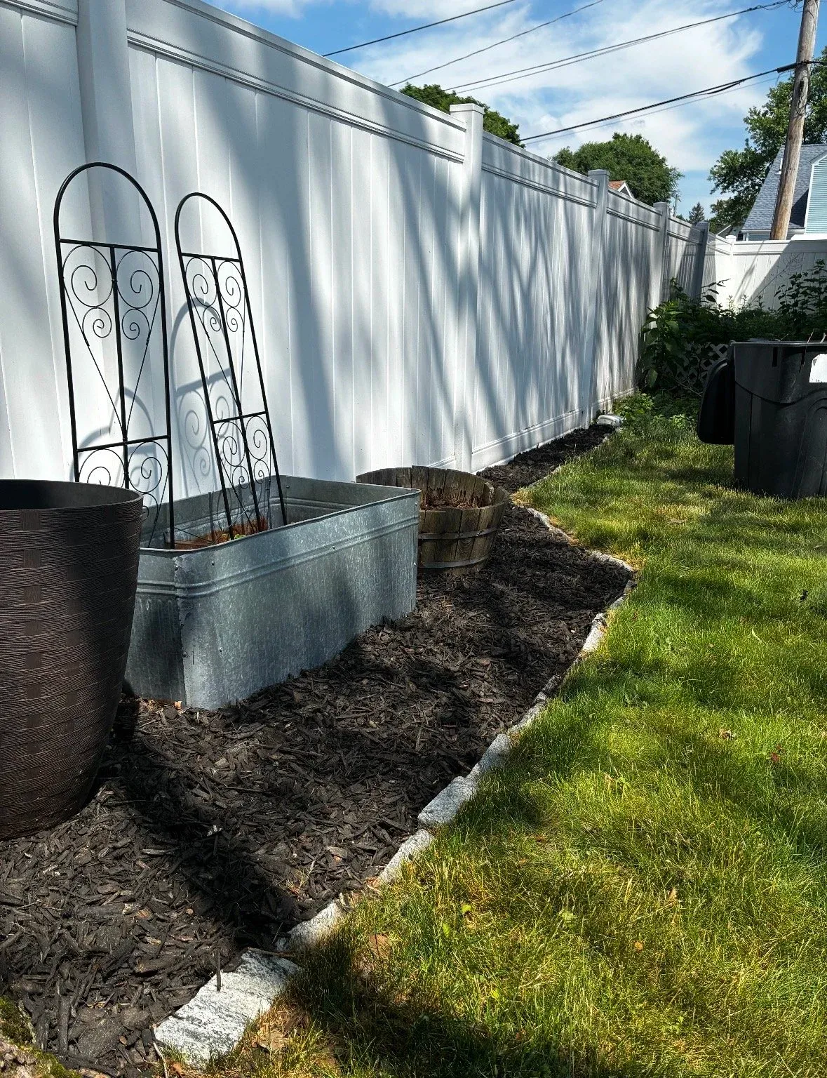 A backyard with a white vinyl fence, two metal plant supports, a galvanized metal planter, a wooden barrel planter, and trash cans. The ground has a mulched garden bed next to a grassy lawn, with shadows of tree branches on the fence.