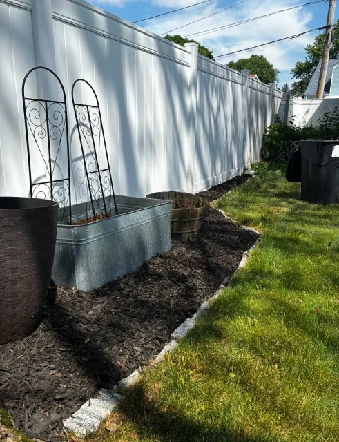 A backyard with a white vinyl fence, two metal plant supports, a galvanized metal planter, a wooden barrel planter, and trash cans. The ground has a mulched garden bed next to a grassy lawn, with shadows of tree branches on the fence.