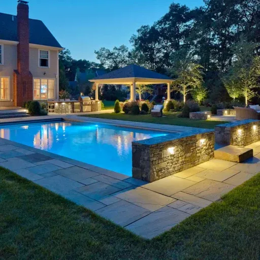 This Sudbury pool landscape uses stone walls, exterior lighting, and layered outdoor living spaces to create a polished evening setting.