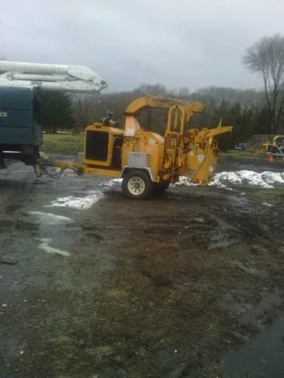 A yellow road construction machine with attached equipment on a muddy, wet ground outdoors on a cloudy day, with trees in the background.