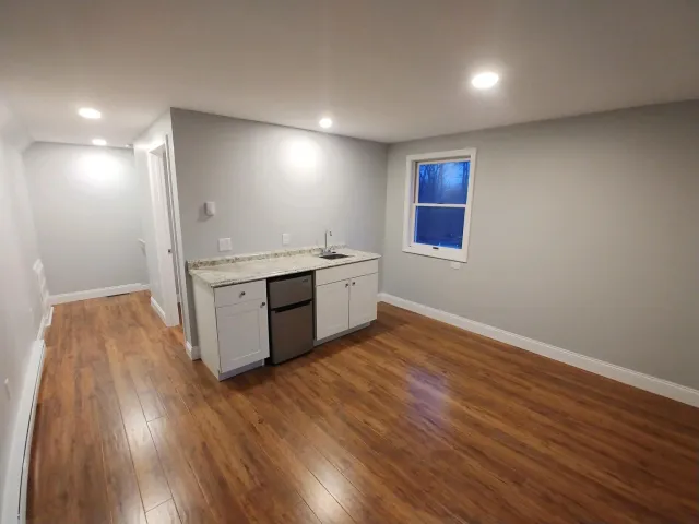 Empty room with wooden floors, a small kitchen area with a granite countertop, white cabinets, a mini refrigerator, a sink, and a window showing a dark outdoor scene.