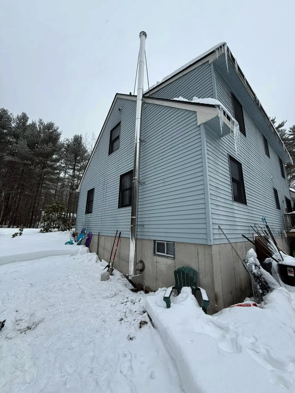A snow-covered house with blue siding and a concrete foundation, surrounded by snow, with icicles hanging from the roof. There are snow shovels, a green chair, and other gardening tools against the house. The background features a snow-covered yard and trees.