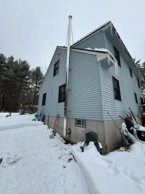 A snow-covered house with blue siding and a concrete foundation, surrounded by snow, with icicles hanging from the roof. There are snow shovels, a green chair, and other gardening tools against the house. The background features a snow-covered yard and trees.