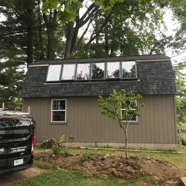 A two-story brown shed with six roof-integrated skylights, a dark shingled roof, and two windows, parked next to a van.
