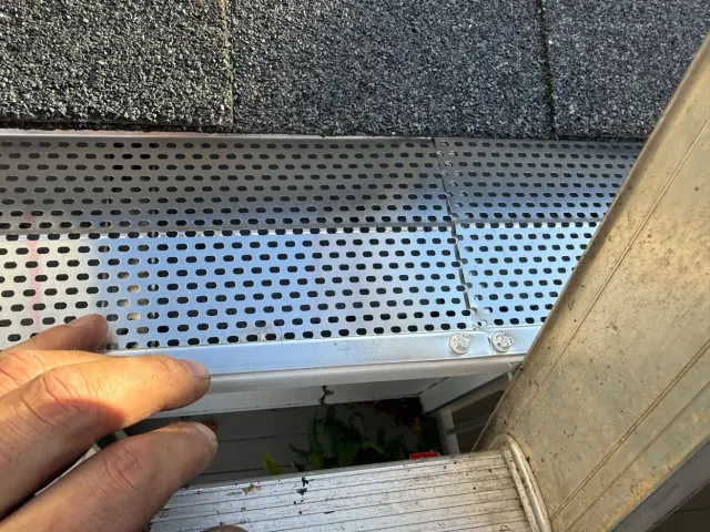 Hand holding a white gutter downspout near a metal roof edge and perforated drain panel
