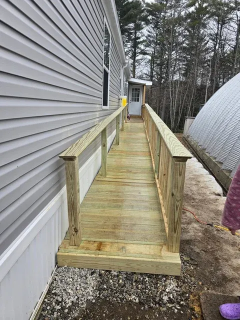 Wooden boardwalk between two buildings with metal siding, bordered by evergreen trees and gravel ground