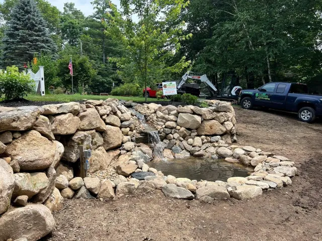 A tiered stone waterfall feature and small pond set in a landscaped yard, with a dark pickup truck parked nearby.
