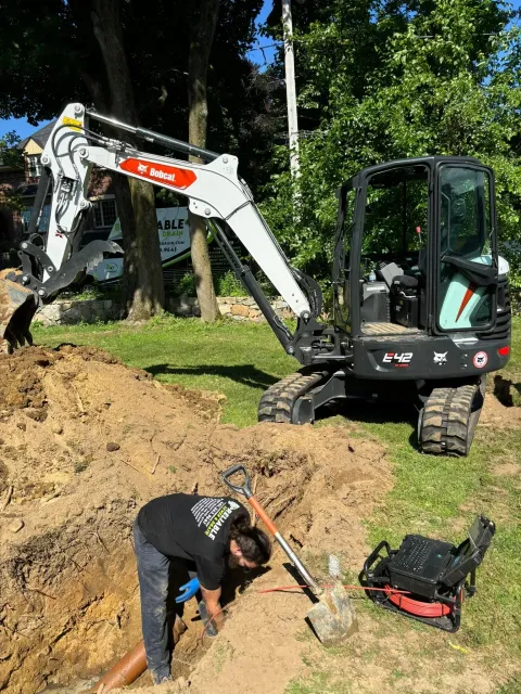 Worker digging near a small excavator at a wooded construction site, with tools and dirt pile nearby