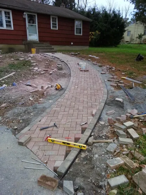 Curved brick walkway under construction beside a red house, with tools and scattered bricks around it