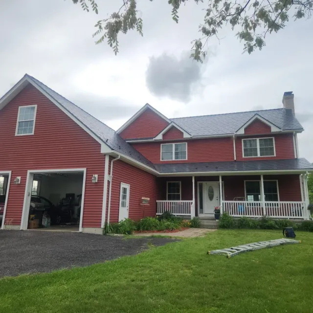 Red farmhouse with attached garage, white porch, and green lawn under a cloudy sky.