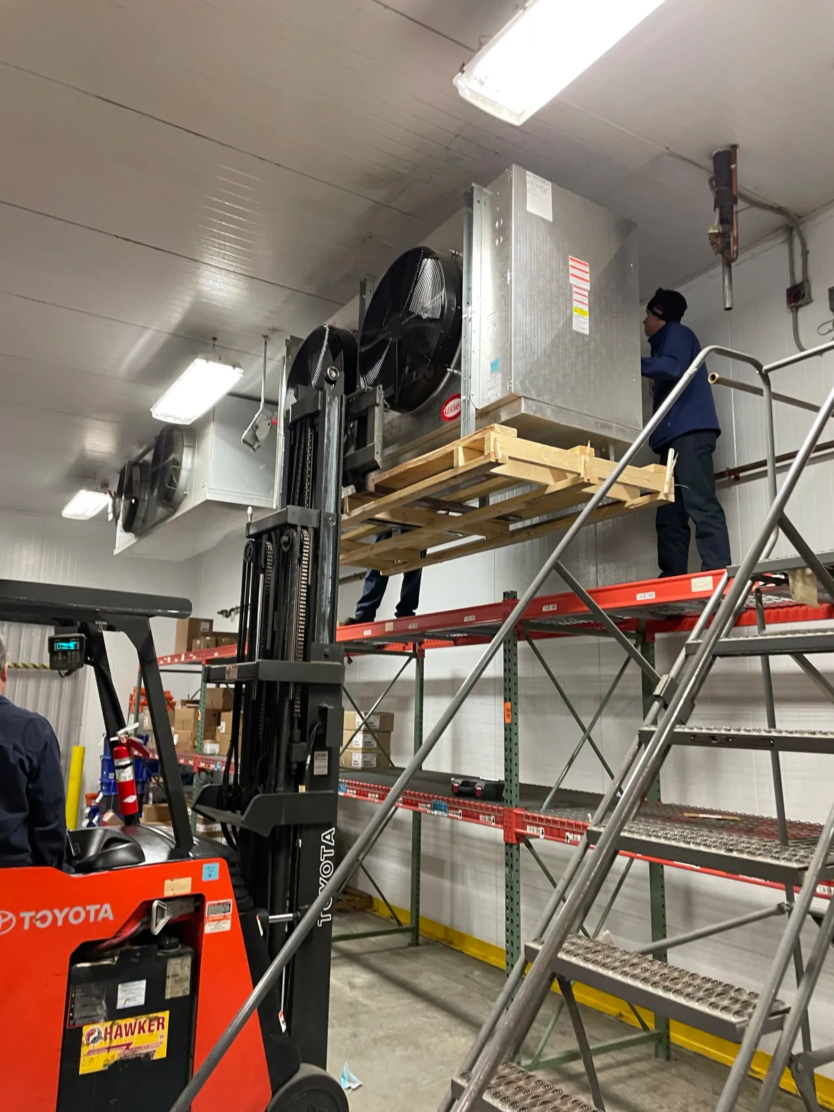 Warehouse setting with a red Toyota forklift, elevated pallets, and workers installing or maintaining industrial equipment on shelves.