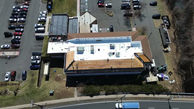 Aerial view of a building with a flat roof, some areas under construction, adjacent parking lot with cars, surrounding trees, and a curved road.
