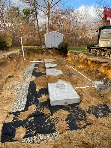 Septic system installation in progress: Open trench with concrete tanks, gravel, and black fabric in a yard.