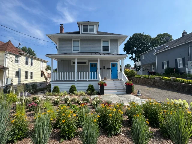 A two-story house with gray siding, blue doors, and a front porch. There are flower beds with yellow and pink flowers, shrubbery, and a stone pathway leading to the house. The sky is clear with a few clouds.
