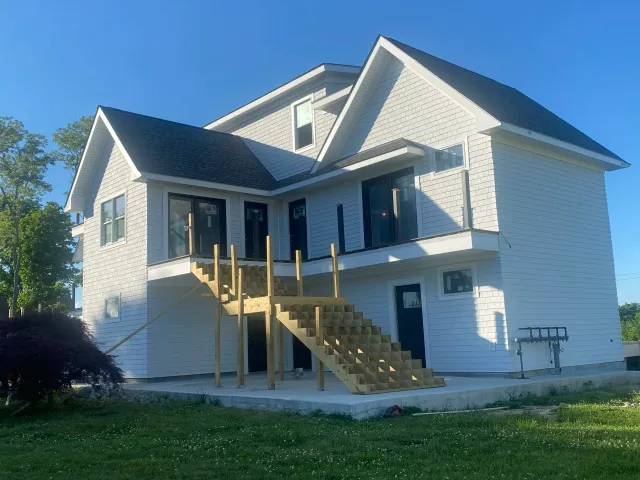 A two-story house under construction with wooden stairs leading to a balcony. The house has white siding, multiple windows, and a dark gray roof. The front yard has green grass and a small tree.
