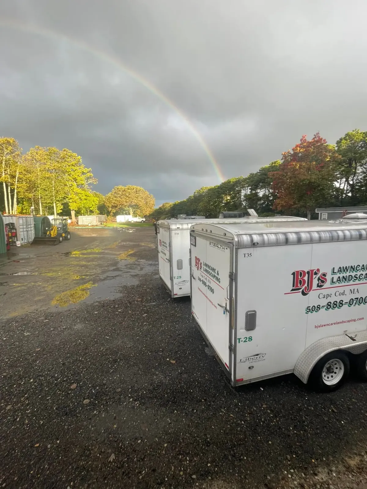 Two white trailers parked on a gravel lot under a rainbow with trees and overcast sky.