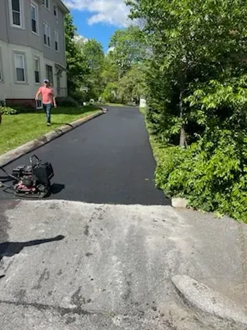 Freshly paved asphalt driveway. A man walks away in the background.  A vibrating compactor sits on the left.