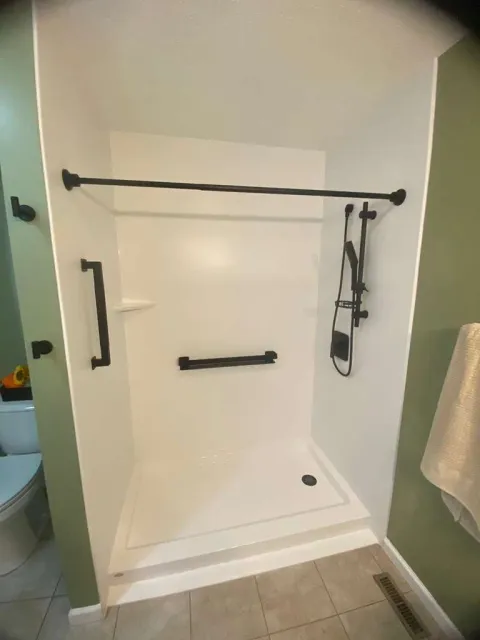 Empty walk-in shower with white walls, black grab bars, a showerhead with a hose, and a corner shelf, next to a green bathroom wall and tile floor.