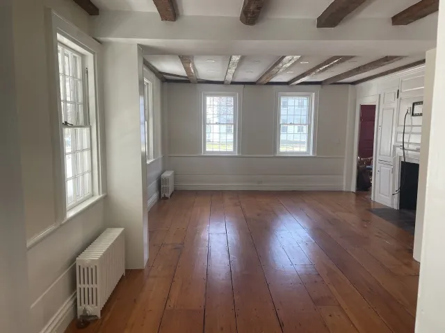 Empty living room with hardwood floor, white walls, and multiple large windows. Exposed wooden ceiling beams add rustic charm. Partially visible adjacent room with maroon wall.