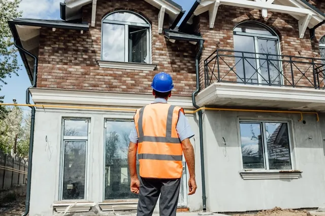 A professional wearing a blue hard hat and high-visibility vest looks up at a two-story brick house while providing home inspection services.