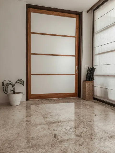 Image of a door with wooden frame and frosted glass panels, positioned against a white wall, with a potted plant on the left and a container holding rifles on the right, near a window covered with a white blind.