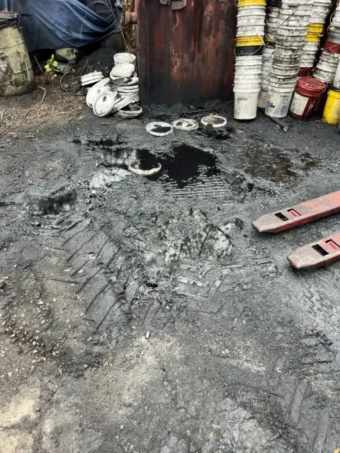 A large oil spill on asphalt, surrounded by stacks of plastic buckets and pallet jack forks.