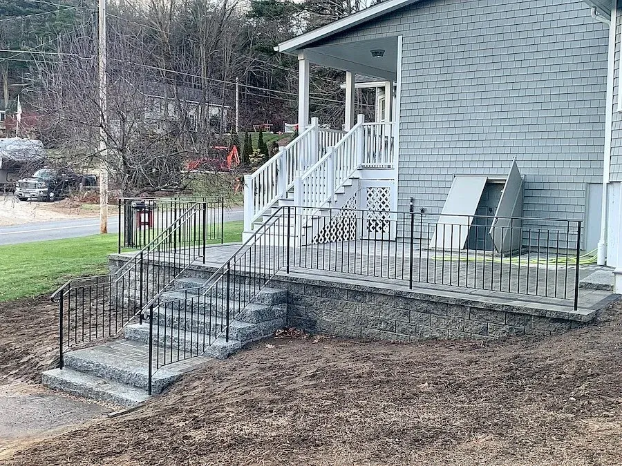 Concrete steps with black metal railings leading to a house porch with white railing and stairs, gray siding house exterior, and an outdoor storage box.