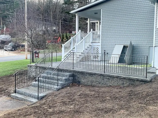 Concrete steps with black metal railings leading to a house porch with white railing and stairs, gray siding house exterior, and an outdoor storage box.