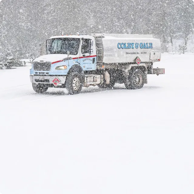 Heavy snow falls around a large silver oil truck with painted light blue letters spelling Colby & Gale