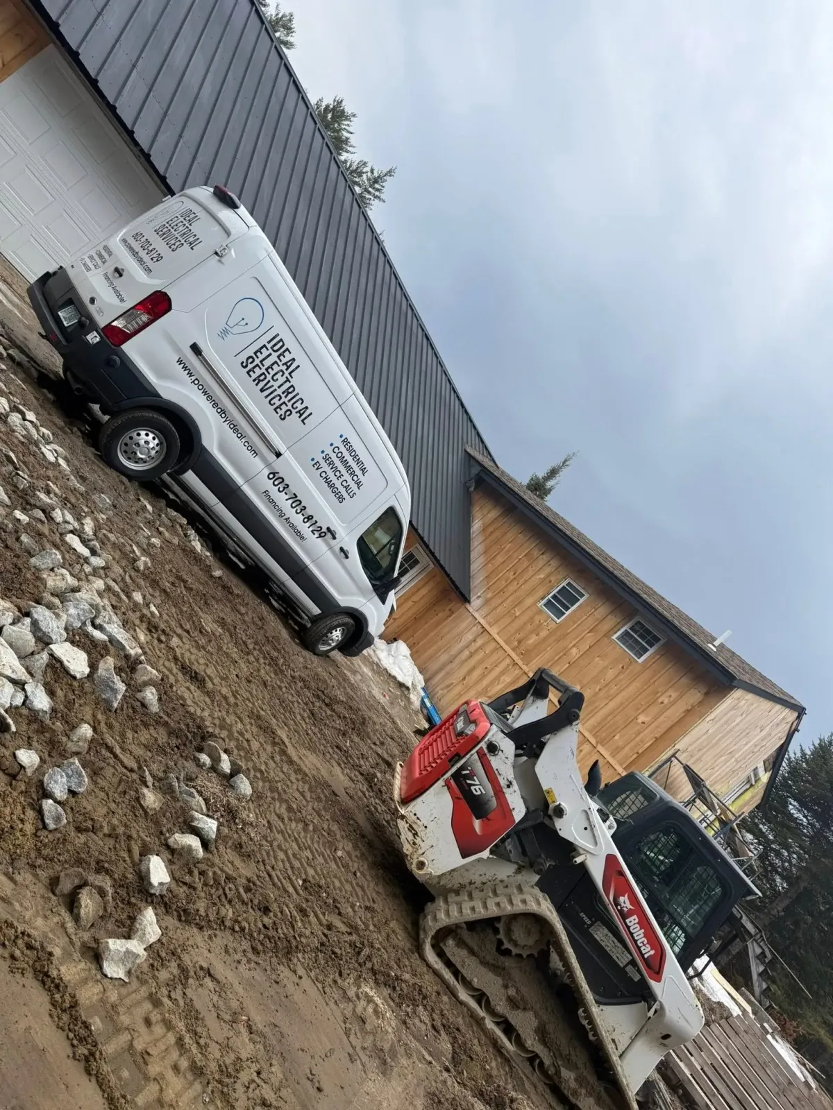 White work van and skid steer on muddy construction site beside a wooden building.