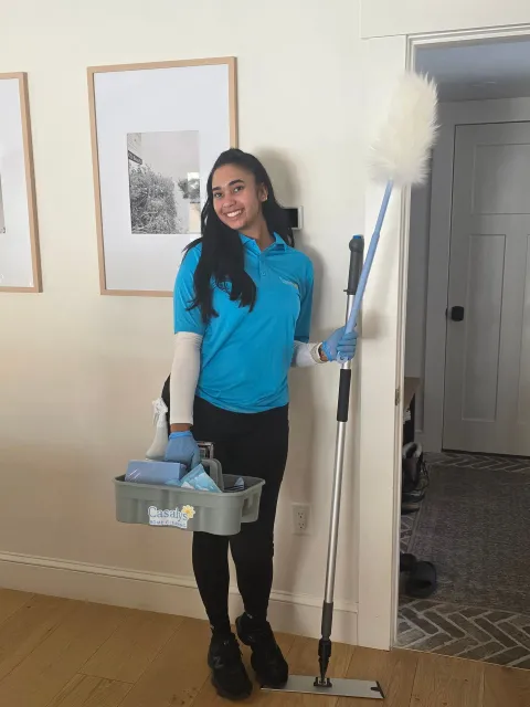 Woman in blue shirt holding cleaning supplies and a mop beside a doorway in a room.