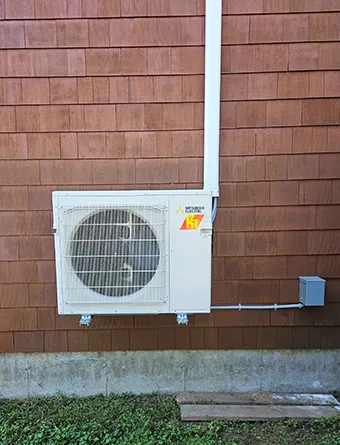 Mitsubishi hyper heat pump is mounted on the exterior wall of a brown shingled building with a conduit running upward, installed by Endless Energy, a Mitsubishi Diamond Contractor