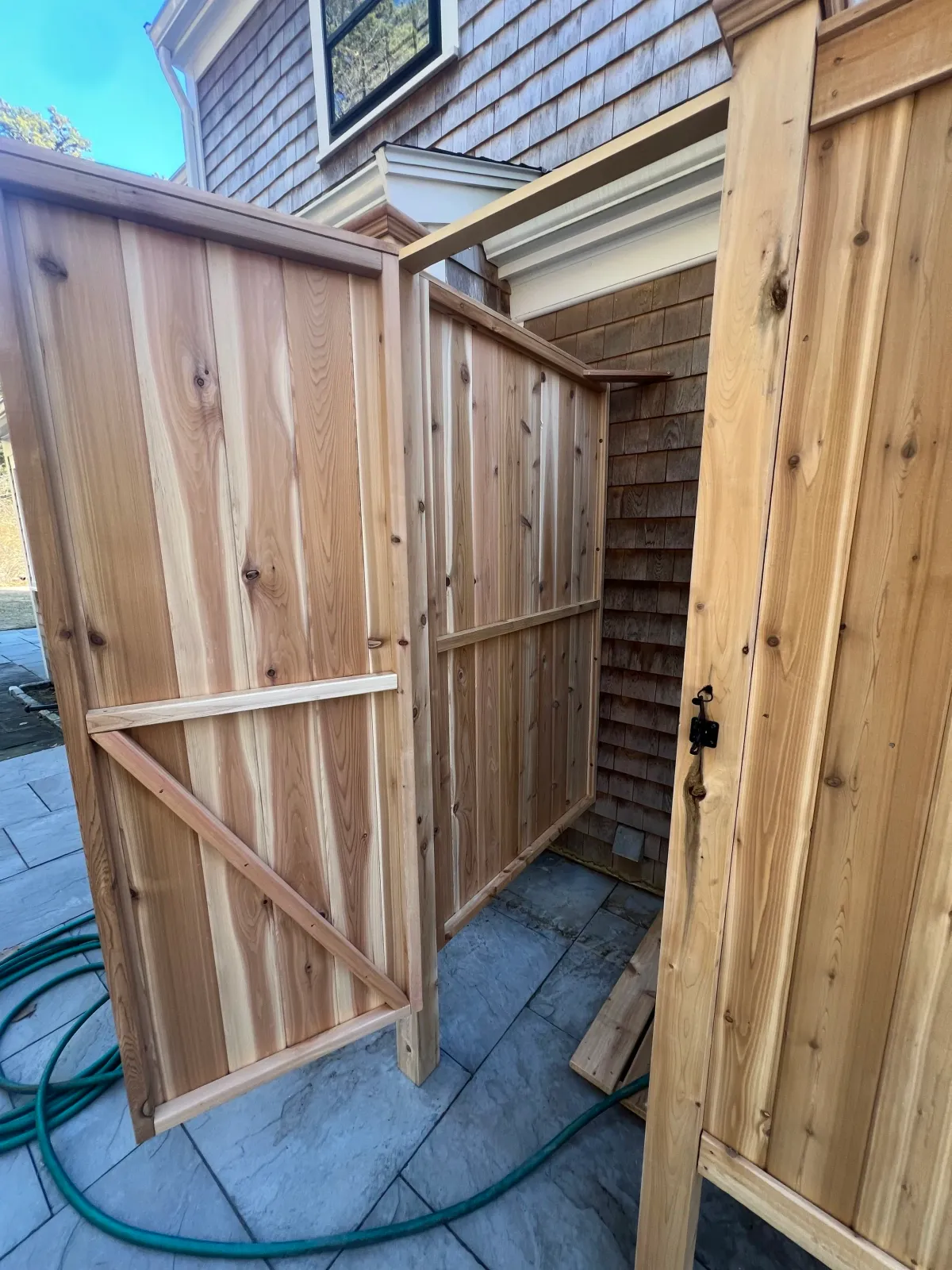 Unfinished outdoor wooden shower or privacy stall with a door lock, on a stone patio with garden hose nearby.