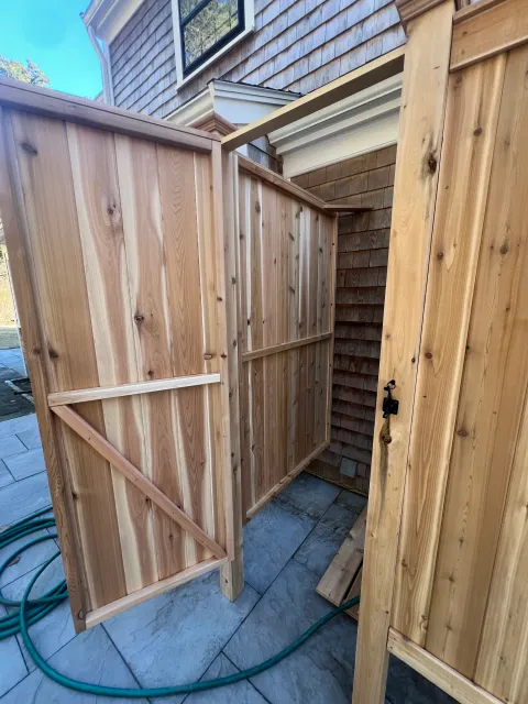 Unfinished outdoor wooden shower or privacy stall with a door lock, on a stone patio with garden hose nearby.
