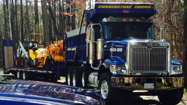 Blue dump truck hauling orange construction equipment on a wooded roadside with another vehicle in the foreground