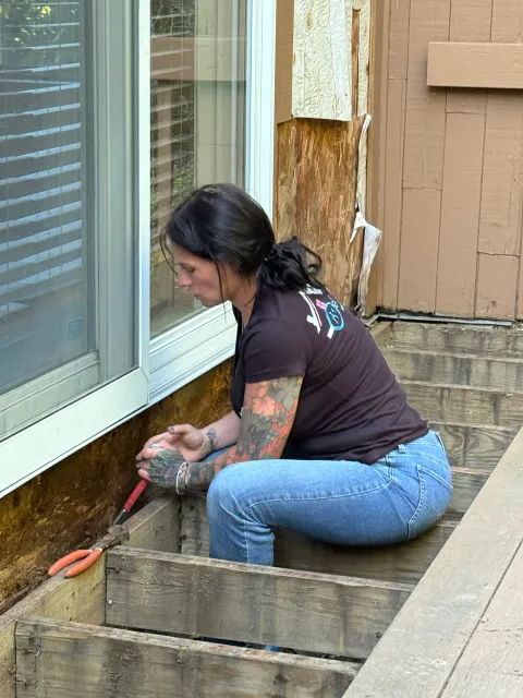 A woman with tattoos on her arms is kneeling on a wooden deck, working with a small tool near the base of a house siding and window.