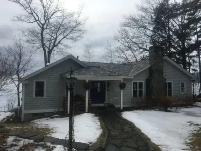 A one-story light gray house with a stone chimney and walkway, surrounded by snow and bare trees.