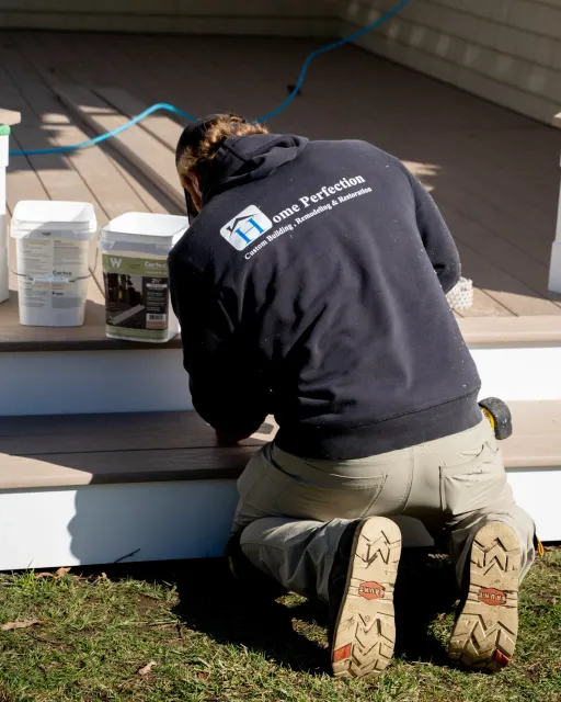A person kneeling on the ground working on a deck, wearing a black hoodie with the words 'Home Perfection' and a logo on the back, surrounded by buckets of paint or stain.