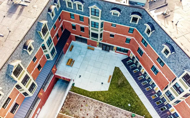 An aerial view of a courtyard surrounded by a red brick residential building with gray roofs, dormer windows, and benches. The courtyard has a concrete patio area, some grassy patches, and a small tree.