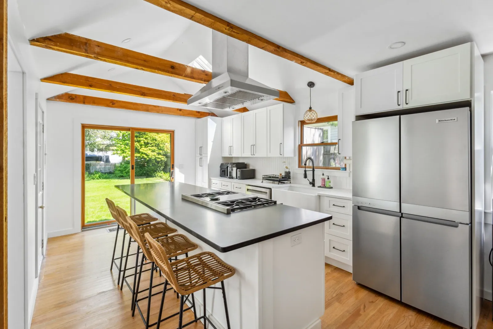 Modern kitchen with white cabinets, stainless steel refrigerator, wooden ceiling beams, a central island with four wicker chairs, and sliding glass doors opening to a green backyard.