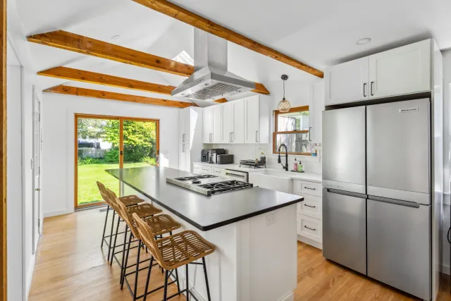 Modern kitchen with white cabinets, stainless steel refrigerator, wooden ceiling beams, a central island with four wicker chairs, and sliding glass doors opening to a green backyard.