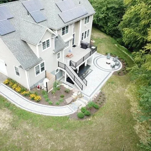 A bird's-eye view of a backyard patio area attached to a multi-story house, featuring a patio with outdoor seating, a staircase leading down to a landscaped yard with flowers and shrubs, and surrounded by trees.