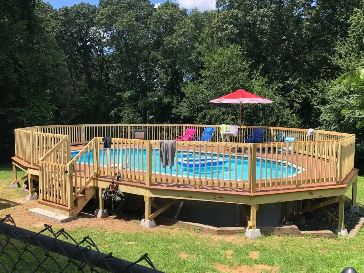 Above-ground pool with a wooden deck and fence, umbrella, and chairs in a grassy yard.