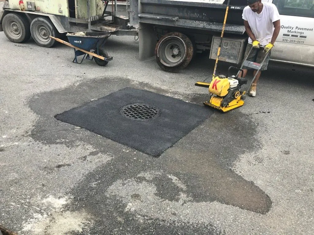 Worker operating a yellow road roller over a black asphalt patch on a street beside a truck