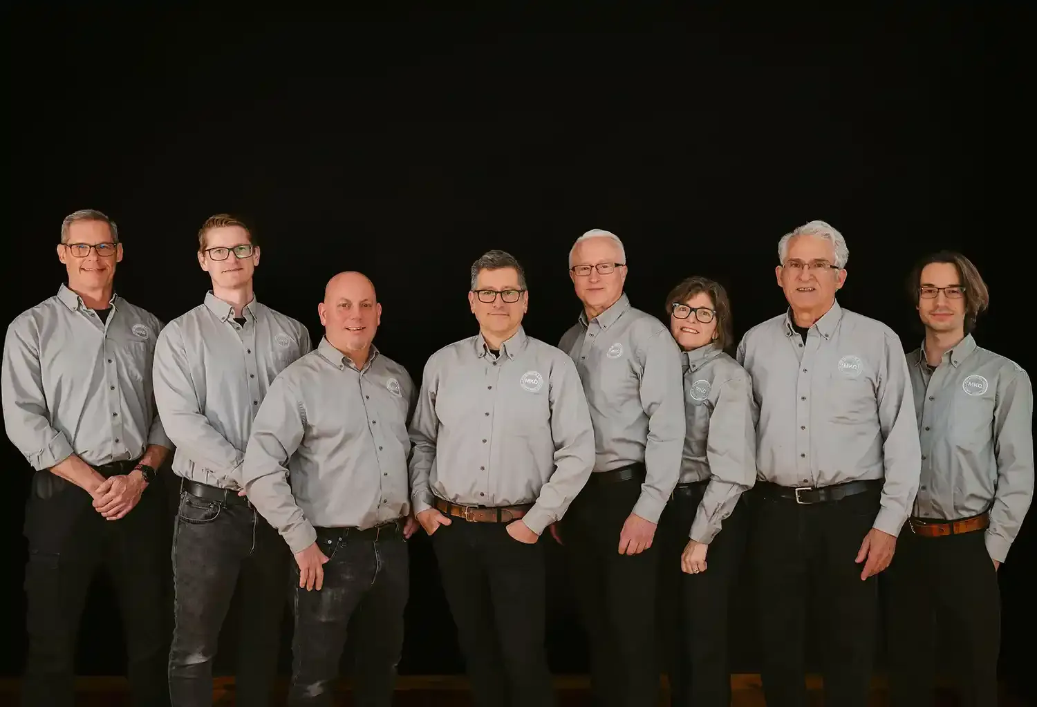Group of eight people standing in a row wearing matching gray button-up shirts with a circular logo on the chest, against a black background.
