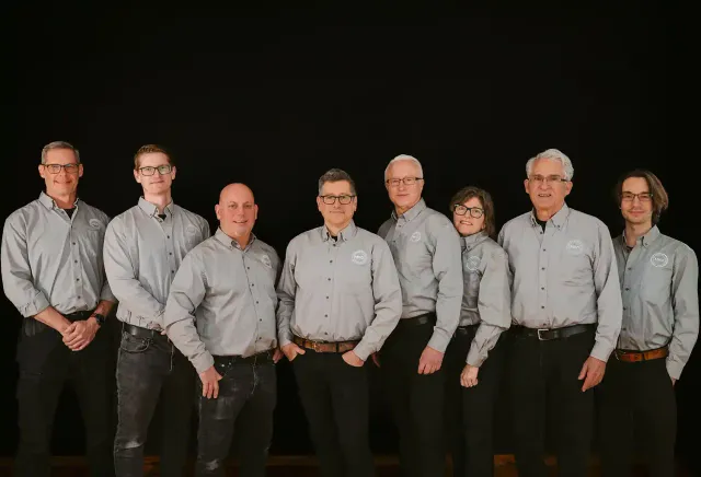 Group of eight people standing in a row wearing matching gray button-up shirts with a circular logo on the chest, against a black background.