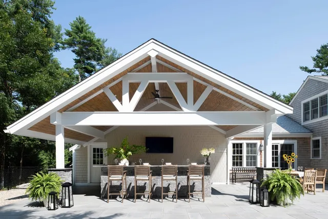 Modern patio with gabled roof, TV, potted plants, lanterns—bordered by trees. – Woodland Builders, South Shore MA.